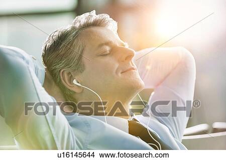 Picture - Mature businessman sleeping while listening to music with earphones on in airport with lens flare. Fotosearch