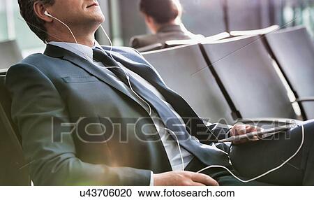 Stock Image - Mature businessman sleeping while listening to music with earphones on in airport with lens flare. Fotosearch