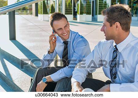 Mature businessman smiling and talking on smartphone while sitting with colleague View Large Photo Image Stock Image - Mature businessman smiling and talking on smartphone while sitting with colleague. Fotosearch