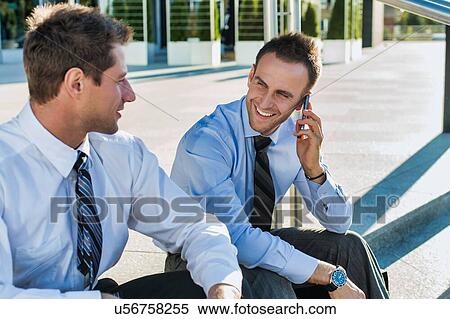 Stock Photography - Mature businessman smiling and talking on smartphone while sitting with colleague. Fotosearch