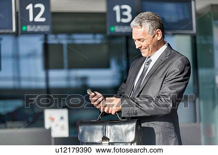 Stock Image - Mature businessman smiling while using smartphone in airport. Fotosearch