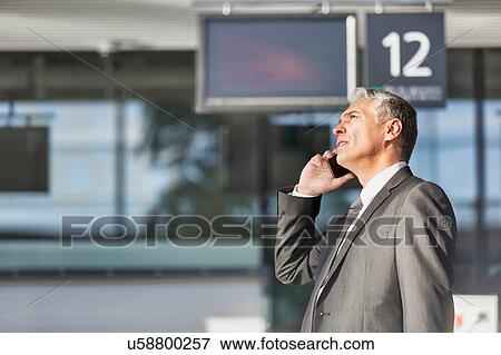 Stock Photo - Mature businessman talking on smartphone while standing at boarding in airport. Fotosearch