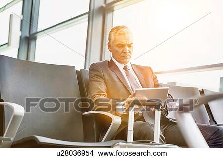Picture - Mature businessman using digital tablet while sitting and waiting for boarding in his gate at airport. Fotosearch