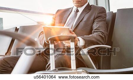 Picture - Mature businessman using digital tablet while sitting and waiting for boarding in his gate at airport. Fotosearch