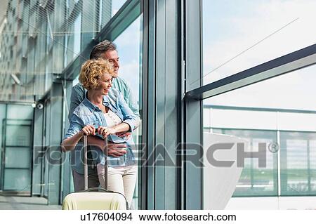 Picture - Mature man hugging his wife from behind while looking through the window in airport. Fotosearch