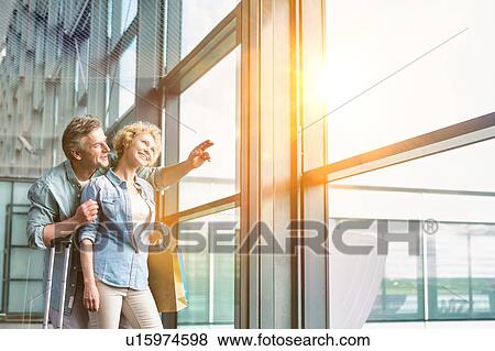 Stock Photo - Mature man hugging his wife from behind while looking through the window in airport. Fotosearch