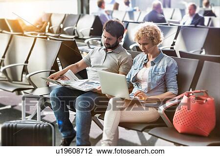 Stock Image - Mature woman working on her laptop, husband is reading newspaper while waiting for boarding in airport. Fotosearch