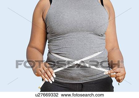 Stock Image - Mid section of woman measuring waist over blue background. Fotosearch