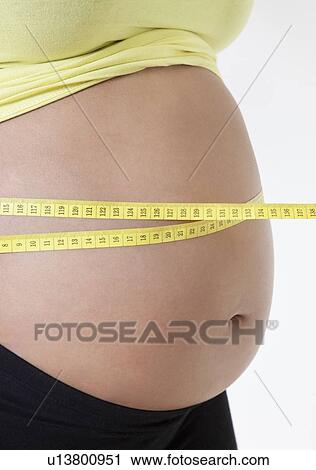 Stock Image - Midsection of pregnant woman measuring stomach with tape against white background. Fotosearch