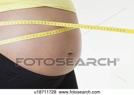 Stock Photo - Midsection of pregnant woman measuring stomach with tape against white background. Fotosearch