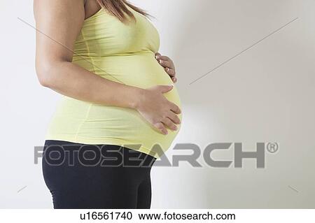 Stock Image - Midsection of young pregnant woman standing against white background. Fotosearch