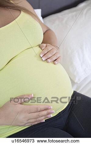 Stock Image - Midsection of young pregnant woman with hands on stomach relaxing at home. Fotosearch