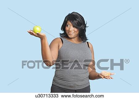 Stock Image - Overweight mixed race woman holding green apple and donut over blue background. Fotosearch