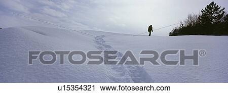 Stock Image - Person walking on snow covered land leaving behind trail of footprints. Fotosearch