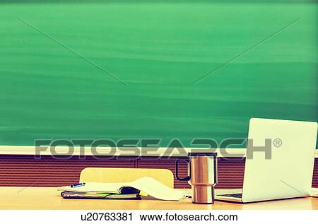 Stock Image - Photo of laptop on professor desk with tumbler and book against black board in classroom. Fotosearch