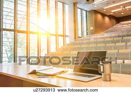 Stock Image - Photo of laptop on professor desk with tumbler in classroom. Fotosearch