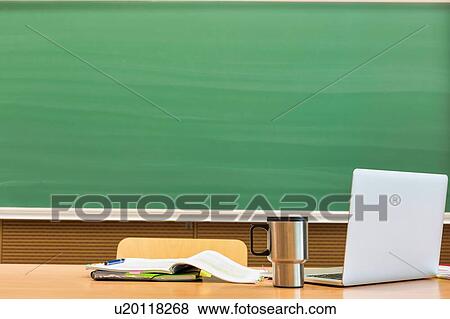 Stock Photo - Photo of laptop on professor desk with tumbler and book against black board in classroom. Fotosearch