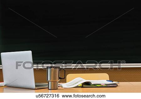 Stock Photograph - Photo of laptop on professor desk with tumbler and book against black board in classroom. Fotosearch