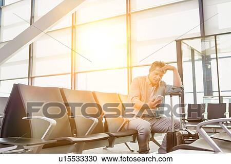 Stock Image - Photo of stressed mature man sitting while looking at his phone in airport. Fotosearch