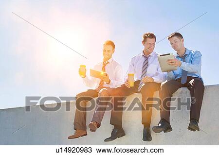 Stock Image - Photo of thoughtful businessmen sitting while using digital tablet in office rooftop . Fotosearch