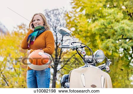 Portrait mature beautiful woman talking on smartphone and holding helmet while standing next to her motorbike in park View Large Photo Image Stock Photo - Portrait mature beautiful woman talking on smartphone and holding helmet while standing next to her motorbike in park. Fotosearch