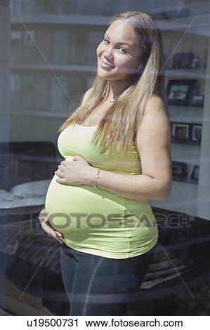 Portrait of a happy pregnant young woman standing with hands on stomach View Large Photo Image Stock Image - Portrait of a happy pregnant young woman standing with hands on stomach. Fotosearch