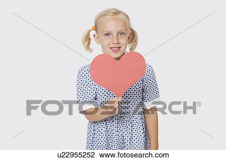 Stock Image - Portrait of a happy young girl holding pink paper heart over white background. Fotosearch