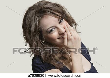 Stock Image - Portrait of a woman smiling with hand over mouth against gray background. Fotosearch