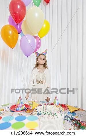 Picture - Portrait of a young girl holding party balloons with cake on table. Fotosearch