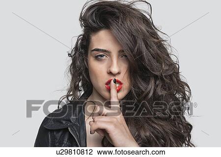 Stock Image - Portrait of a young woman with unkept hair and finger on lips against gray background. Fotosearch