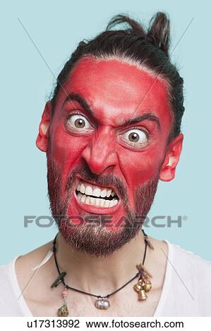 Portrait of aggressive young man with red painted face clenching teeth against blue background View Large Photo Image Stock Image - Portrait of aggressive young man with red painted face clenching teeth against blue background. Fotosearch