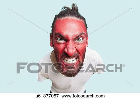 Stock Photo - Portrait of aggressive young man with red painted face clenching teeth against blue background. Fotosearch