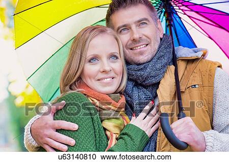 Stock Image - Portrait of attractive man holding umbrella while embracing his wife in park. Fotosearch