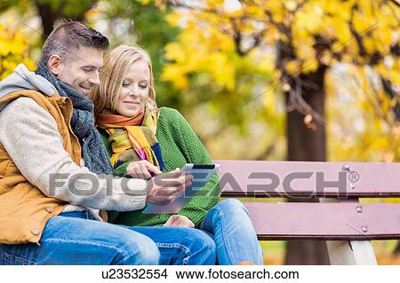Picture - Portrait of attractive man showing digital tablet to his wife while sitting on bench in park. Fotosearch