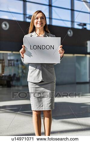 Stock Image - Portrait of attractive woman standing while holding white board with London signage in arrival area at airport. Fotosearch