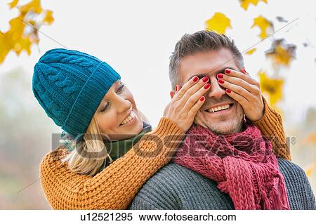 Stock Photography - Portrait of beautiful woman covering her husband eye in park. Fotosearch