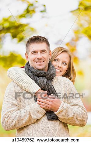 Stock Image - Portrait of beautiful woman hugging her husband from behind in park. Fotosearch