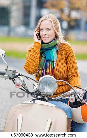 Stock Photo - Portrait of beautiful woman talking on smartphone while waiting and sitting on motorbike in park. Fotosearch