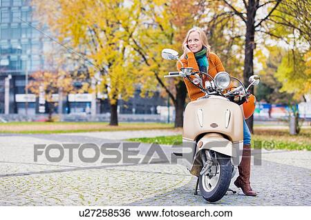 Stock Photograph - Portrait of beautiful woman talking on smartphone while waiting and sitting on motorbike in park. Fotosearch