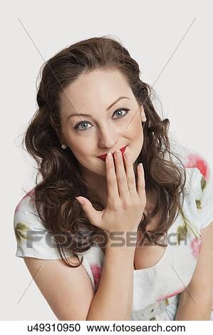 Stock Image - Portrait of beautiful young Caucasian woman smiling with hand over mouth against white background. Fotosearch