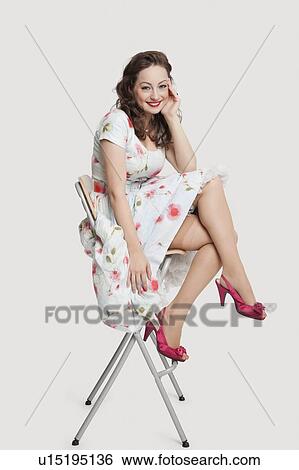 Stock Photograph - Portrait of beautiful young woman in dress sitting on stool against white background. Fotosearch