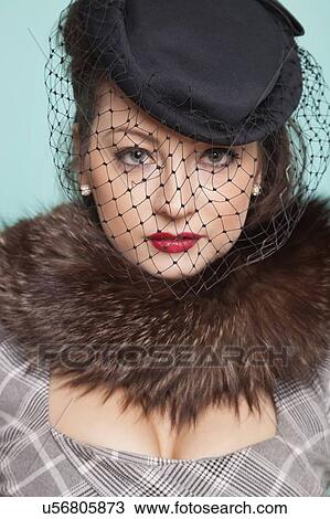 Stock Image - Portrait of beautiful young woman wearing veiled hat with fur boa draped around her neck against blue background. Fotosearch
