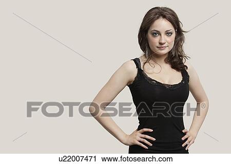 Stock Image - Portrait of beautiful young woman with hands on hip against gray background. Fotosearch