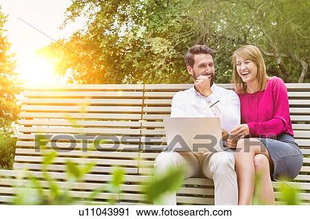 Stock Image - Portrait of businessman and businesswoman sitting on bench while working on laptop. Fotosearch