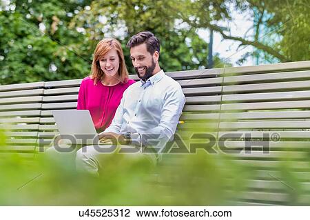 Stock Image - Portrait of businessman and businesswoman sitting on bench while working on laptop. Fotosearch
