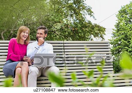 Stock Photo - Portrait of businessman and businesswoman sitting on bench while working on laptop. Fotosearch