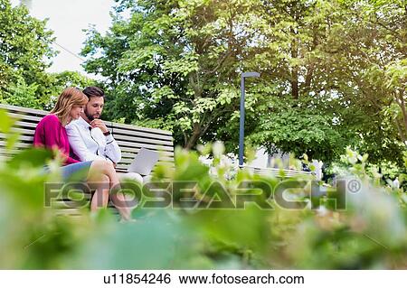 Stock Photograph - Portrait of businessman and businesswoman sitting on bench while working on laptop. Fotosearch