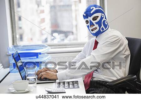 Stock Image - Portrait of businessman using laptop while wearing wrestling mask at office desk. Fotosearch