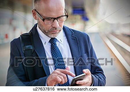 Stock Photograph - Portrait of businessman using smartphone while waiting for the train in station. Fotosearch