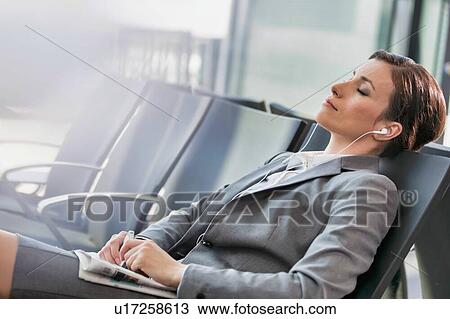 Stock Image - Portrait of businesswoman listening to music on smartphone while sleeping and waiting for boarding in airport. Fotosearch